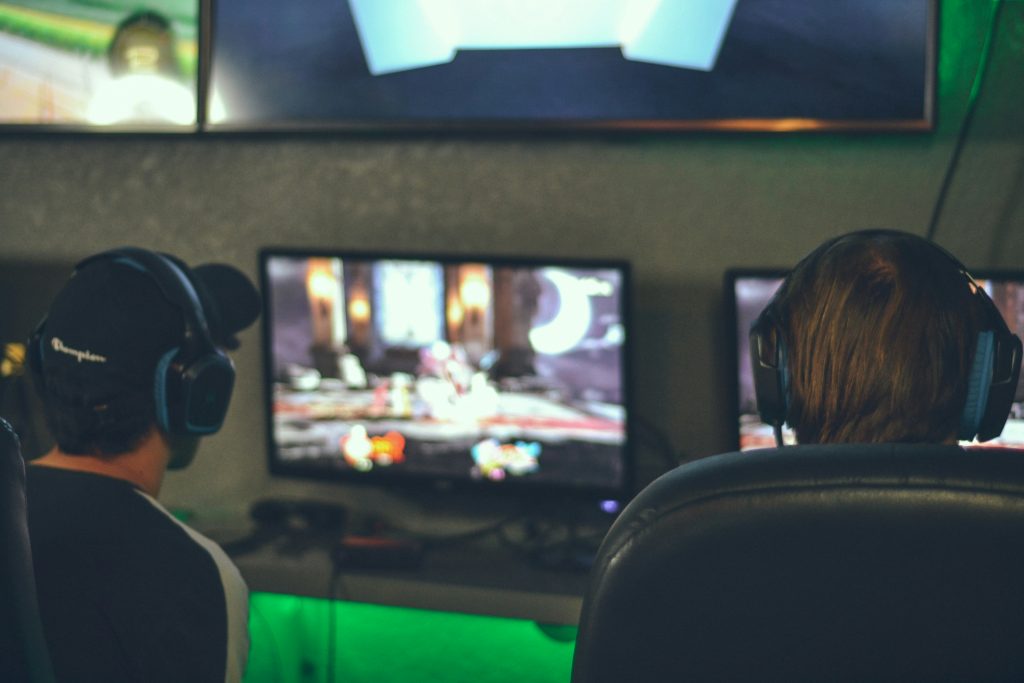 Two people wearing headphones sit side by side gaming at monitors in a dark room with green LED lighting.