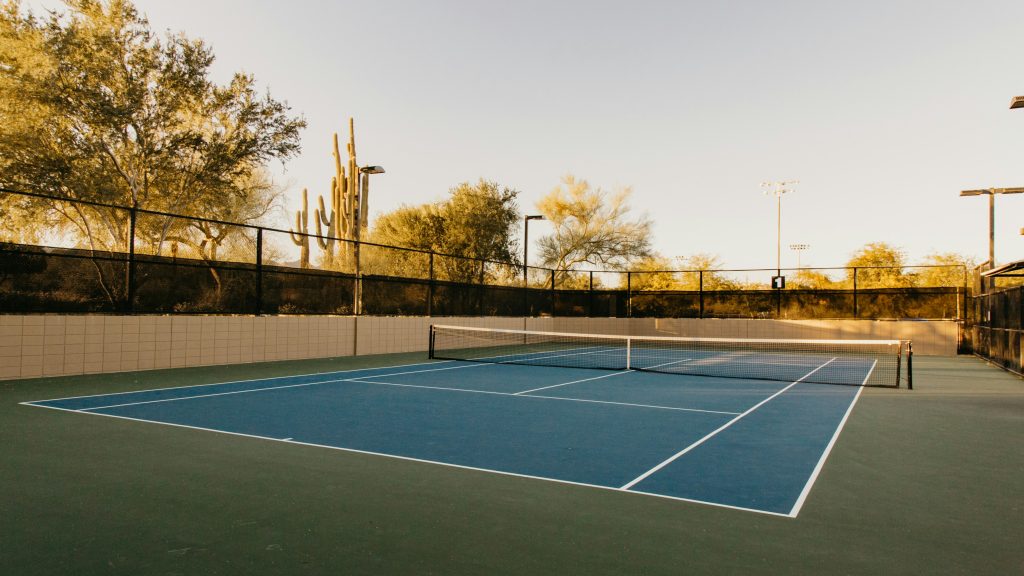 An empty outdoor tennis court with a blue surface, net, and black fence, backed by desert trees and cacti in warm evening light.